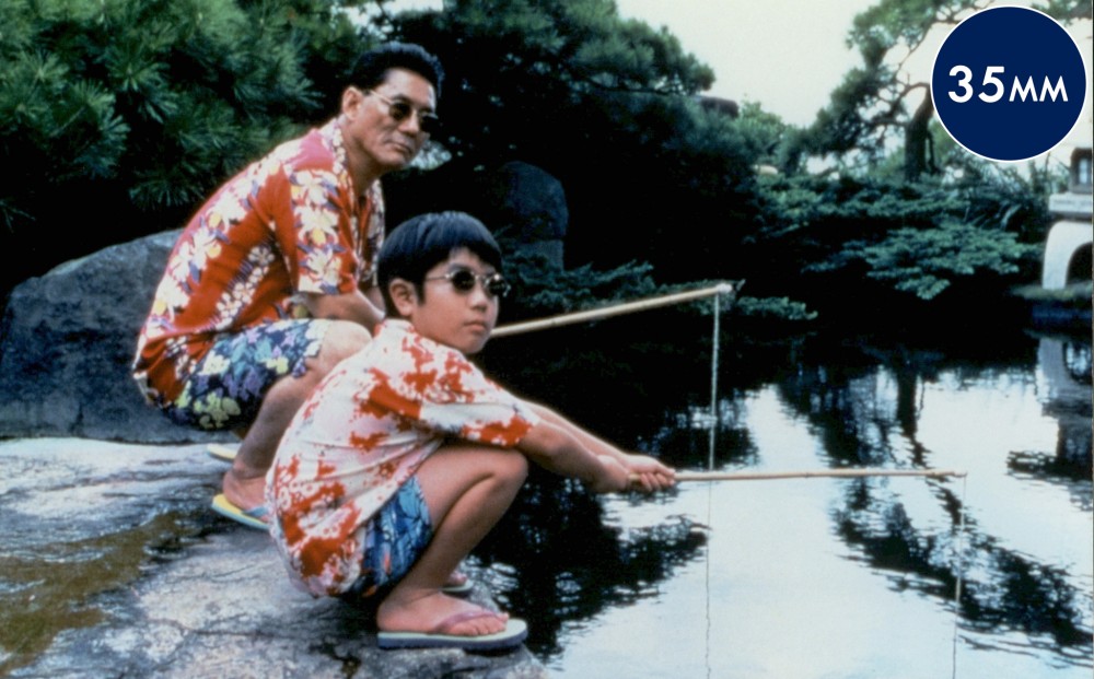 A young boy and a middle-aged man squat by a river, holding fishing poles, and wearing sunglasses, hawaiian shirts, swim trunks, and flip flops.