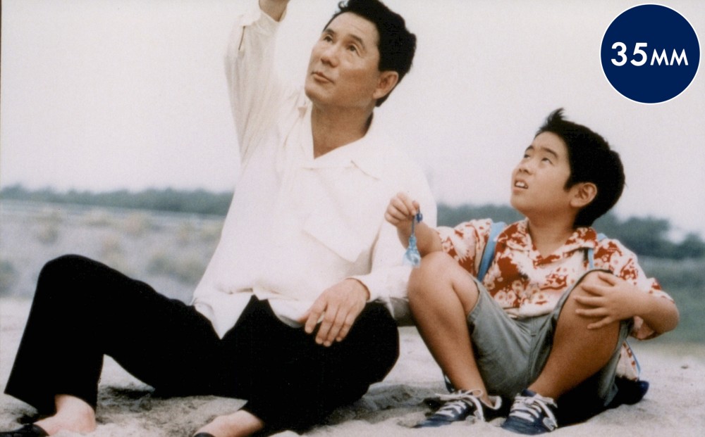 A young boy and a middle-aged man sit next to each other on sand.