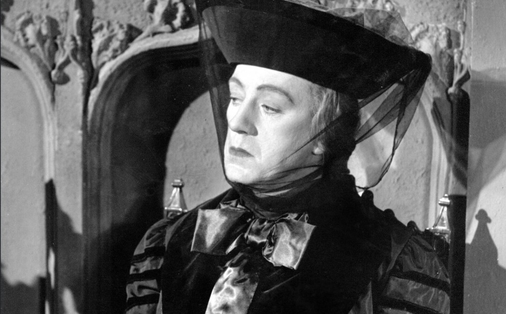 Actor Alec Guinness in character as Lady Agatha D'Ascoyne, wearing a large black hat with a veil.