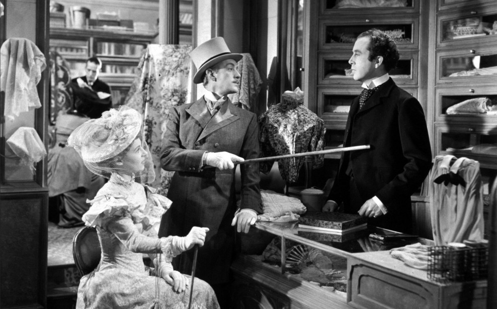 Two people dressed finely address Alec Guinness in the draper's shop where he works.