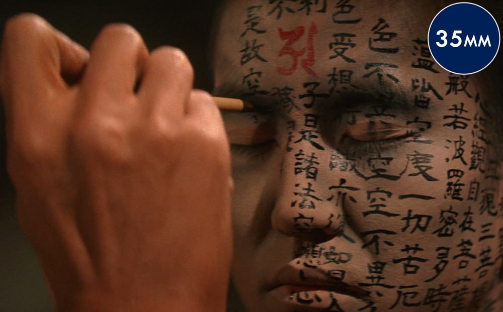 Close-up on a man's face; a hand draws Japanese characters on his face with ink and a brush.