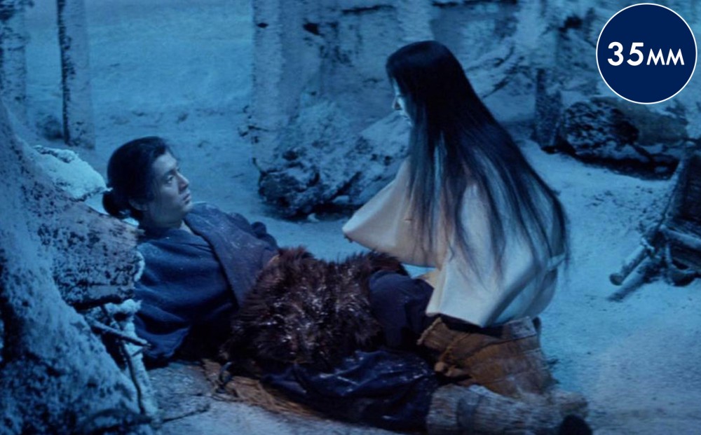 A woman kneels beside a man who is laying on his back in a snowy, blue-lit environment; he gazes at her.