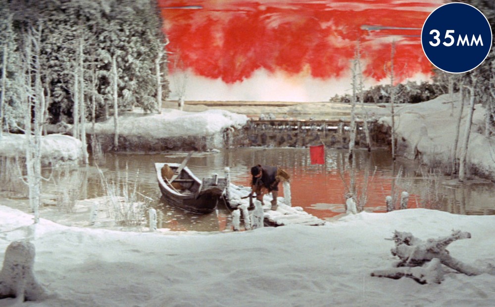 In a snowy landscape, a man bends down on a dock; a canoe is in the water beside the dock.
