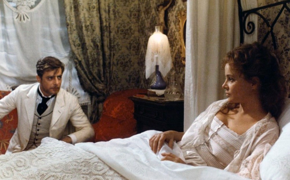 A woman laying in bed and a man seated on a chair by her look at each other; both are dressed in white.