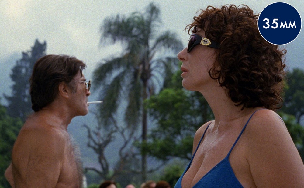 A man and woman stand in profile, each wearing sunglasses and trunks/a swimsuit.