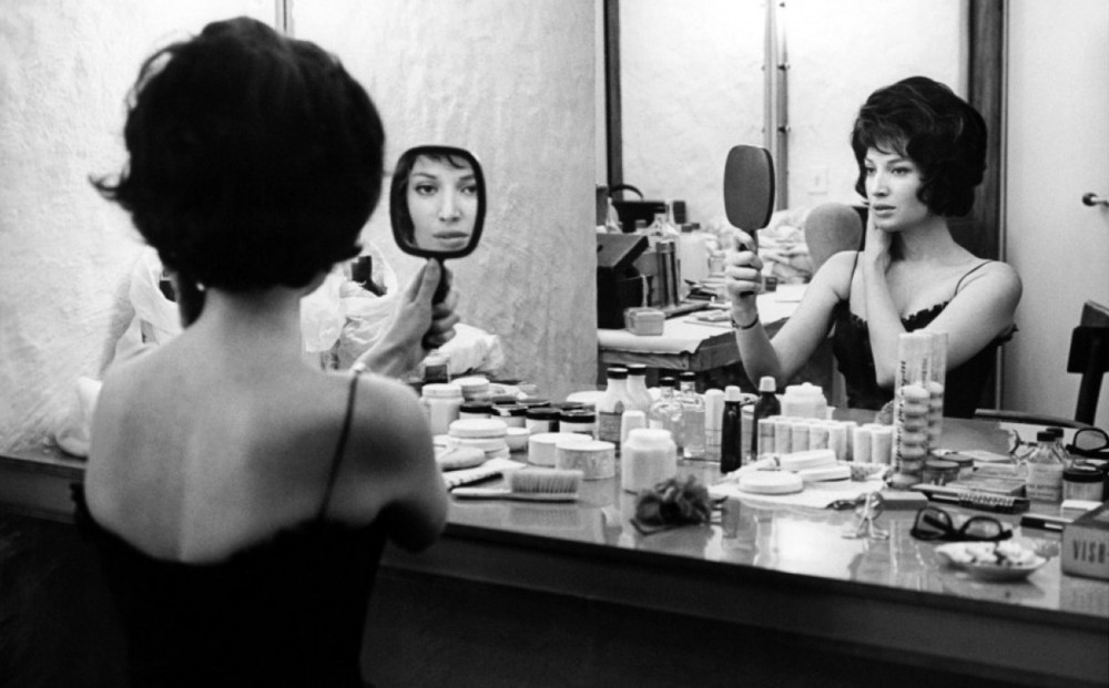 Actor Jeanne Moreau sits at a vanity in front of a large mirror, while also looking in a hand mirror.