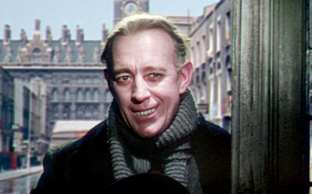 Actor Alec Guinness stands at a doorway, smiling awkwardly.