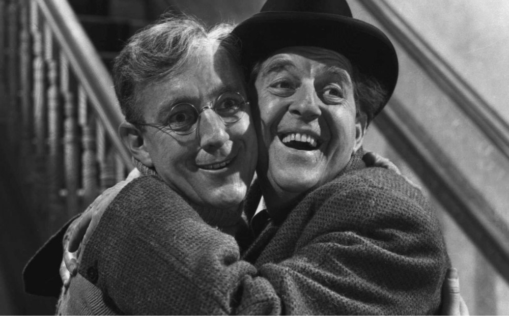 Actors Alec Guinness and Stanley Holloway hug each others, with their smiling faces pressed up against one another; both look out in the same direction.