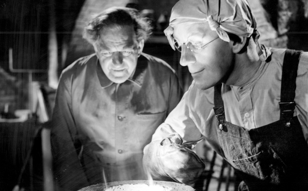 Actors Alec Guinness and Stanley Holloway stand over a vat of shining gold; Guinness wears gloves, work coveralls, gloves, and a little head covering.