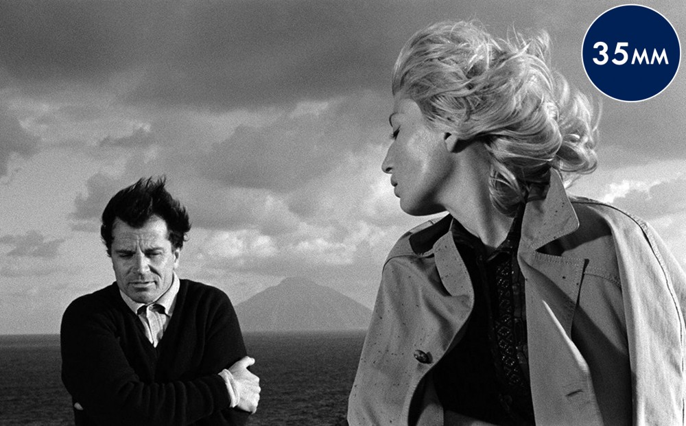 Wind blows the hair of actors Gabriele Ferzetti and Monica Vitti, against a backdrop of the ocean.