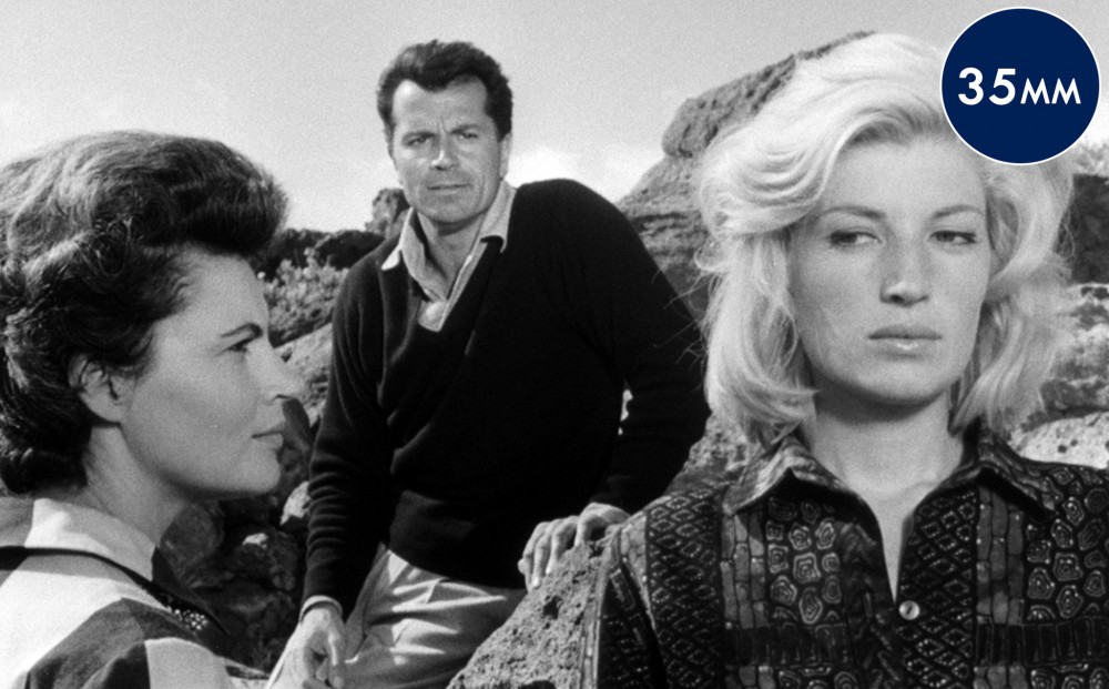 Actor Lea Massari looks at Monica Vitti, who turns her eyes but not her face towards her; actor Gabriele Ferzetti leans on a crop of rocks behind him.