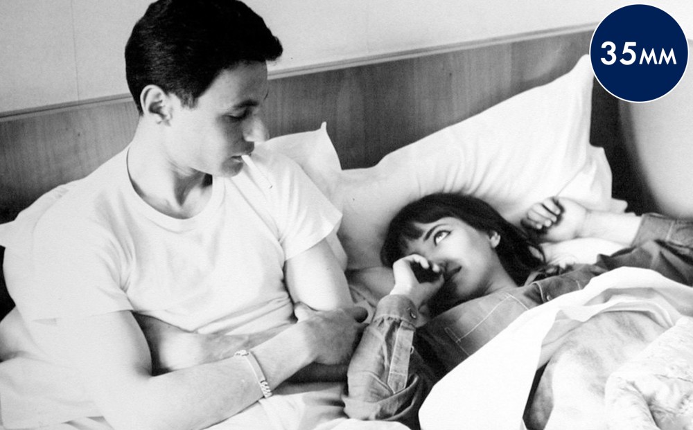 Actor Anna Karina lays in bed, looking up at a man sitting next to her, who looks down at her.