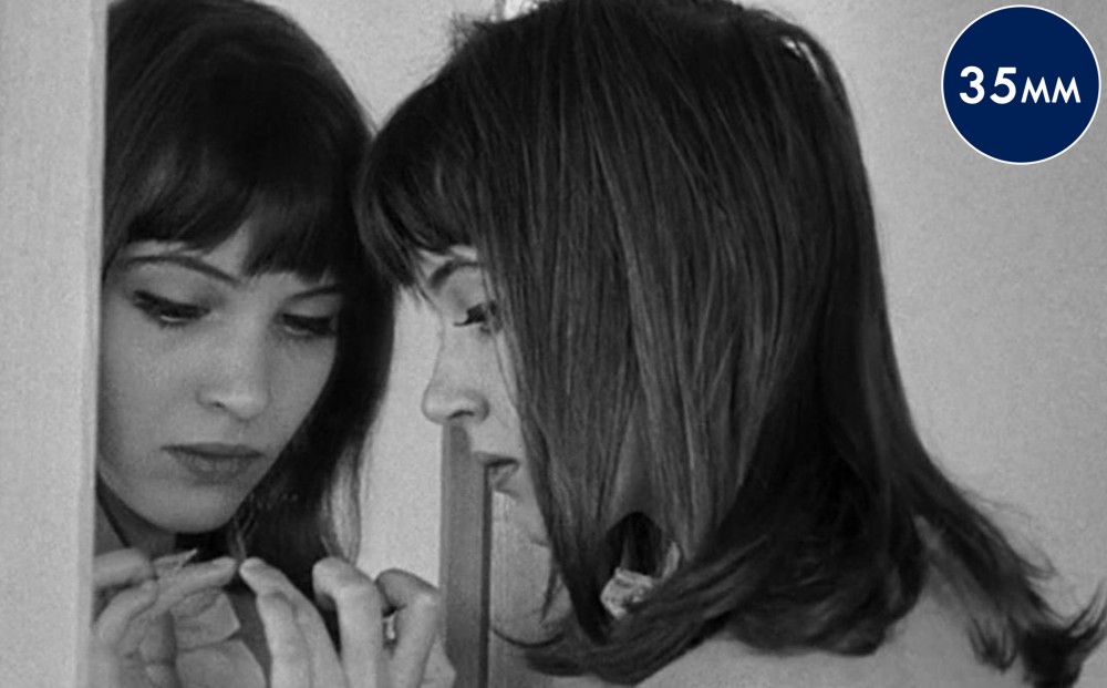 Actor Anna Karina appears in profile and her face can be seen in the mirror that she touches with her fingertips.