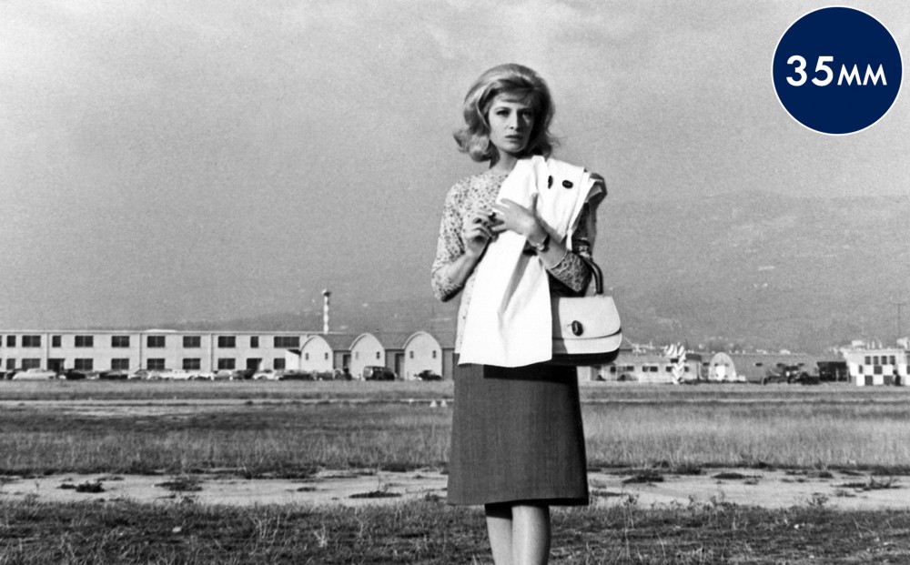 Actor Monica Vitti stands in a barren field, with buildings in the background.