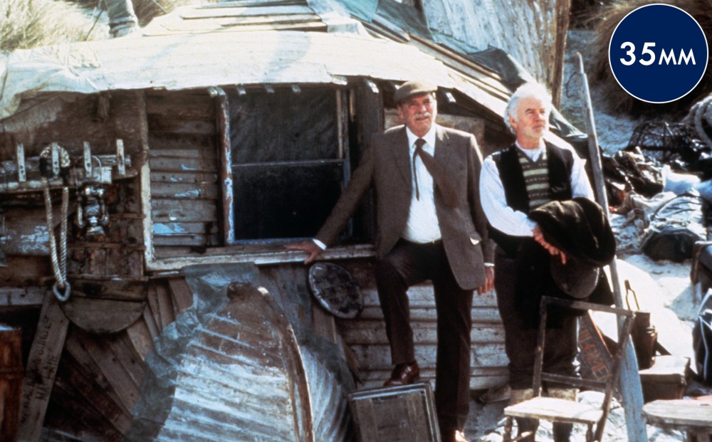 Actor Burt Lancaster and another man stand near a ramshackle beach cottage.