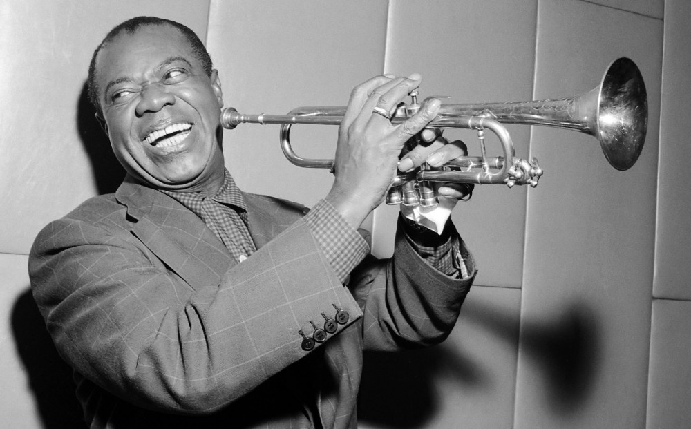 Close-up on Louis Armstrong, who smiles and holds his trumpet.