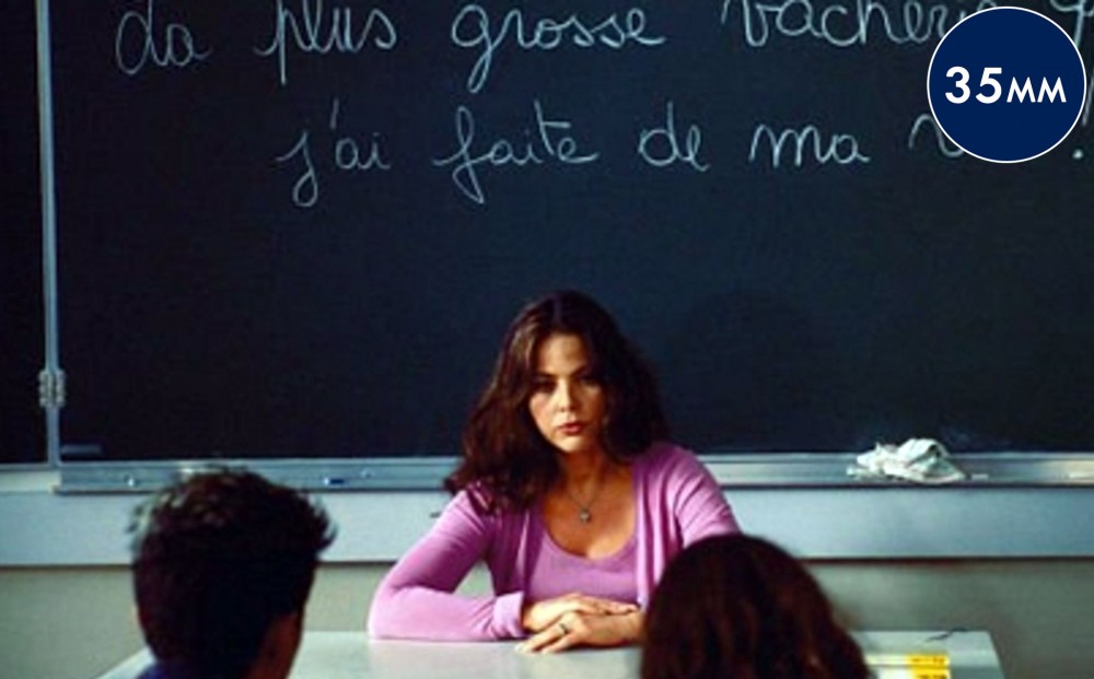 Actor Ornella Muti sits in front of a classroom with a blackboard behind her.