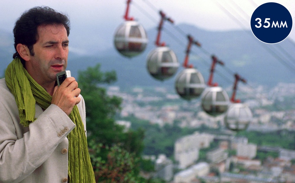 Actor François Morel speaks into a recorder, with an aerial tramway in the background.