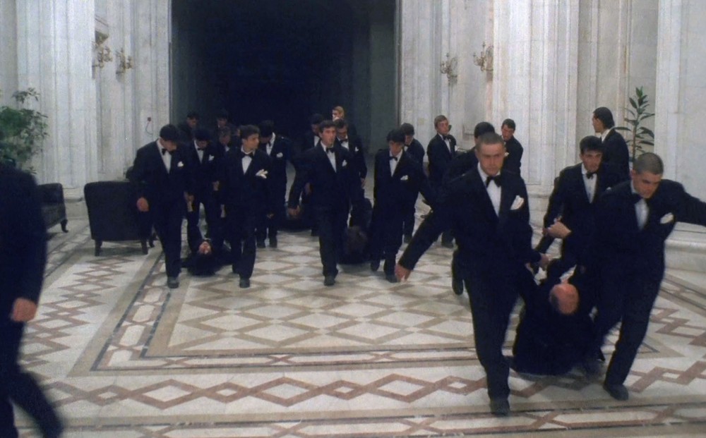 Couples of two men in tuxedos each drag other men in tuxedos through a large, grand looking hall.