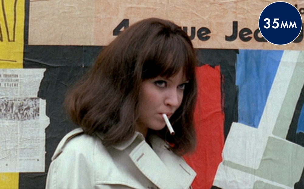 Actor Anna Karina stands by a wall outside, a cigarette in her mouth.