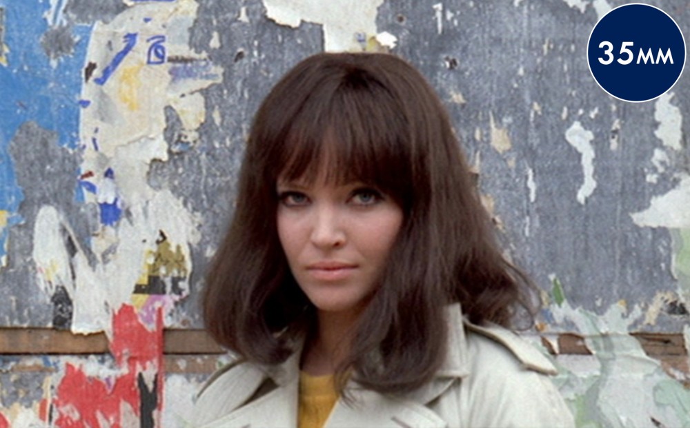 Anna Karina stands outside by a wall, looking towards the camera.