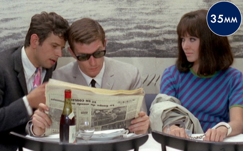 Actor Anna Karina is handcuffed and sits at a dining table next to two men who both look at a newspaper.