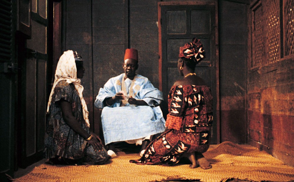 Two women kneel alongside a seated man in a small room.