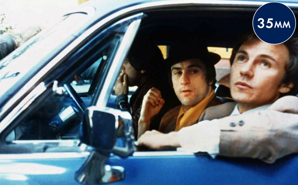 Three men look out of the window of a car; Harvey Keitel in the driver's seat and Robert De Niro in the middle seat.