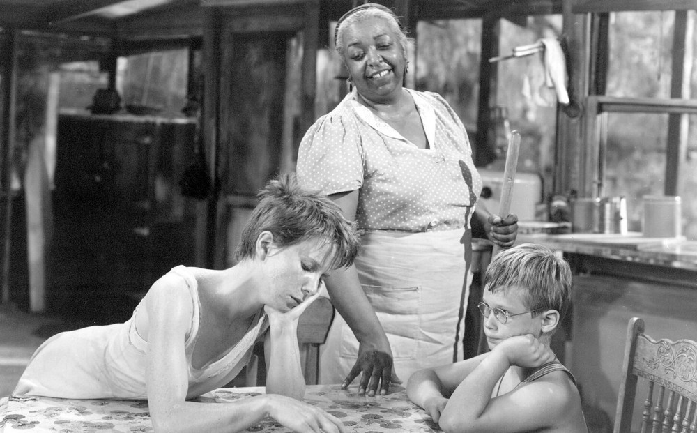 Actor Ethel Waters holds a broom and sets her hand on the table where two young white children are seated.