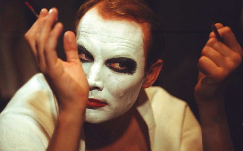 Actor Klaus Maria Brandauer applies makeup, already wearing white face paint and black eyeliner, with red lips.