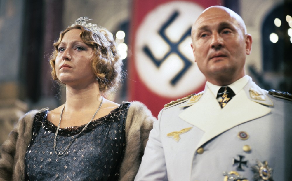 A bald man in a white army officer's uniform and a blonde woman dressed formally stand together, with a Nazi flag in the background.
