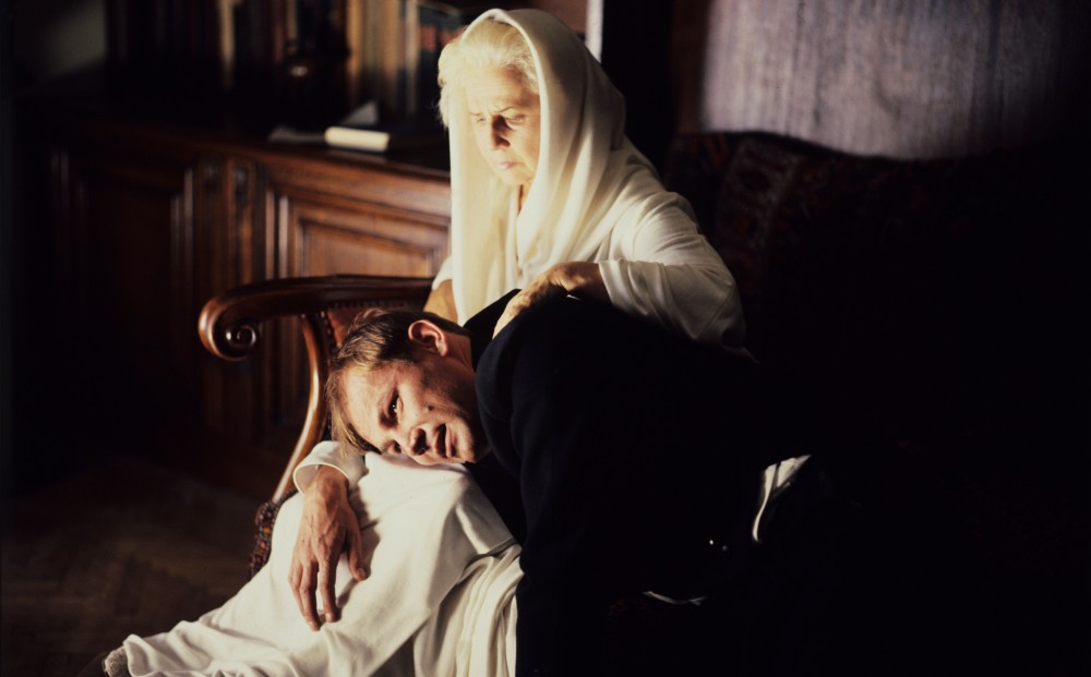 Actor Klaus Maria Brandauer lays his head in the lap of a woman with white hair who is dressed all in white.