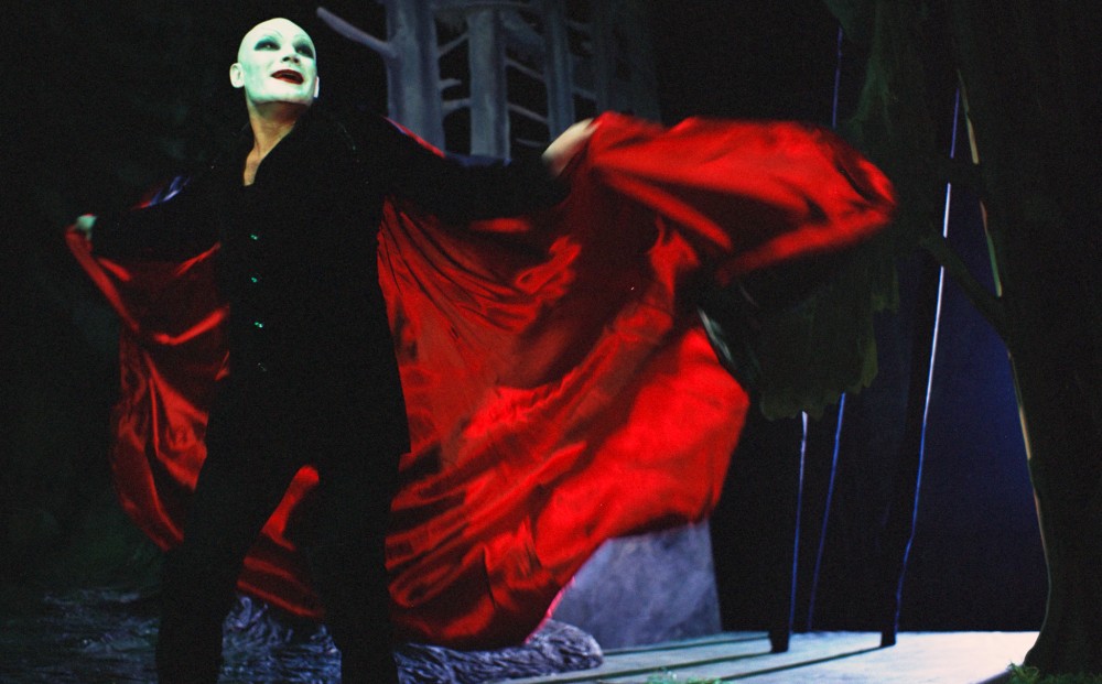 Actor Klaus Maria Brandauer in costume, with white face paint, red lips, a black outfit, and red cape.
