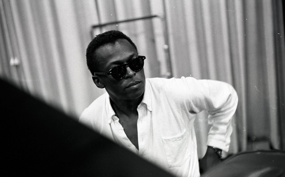 Miles Davis, wearing a white button-down shirt and sunglasses inside.