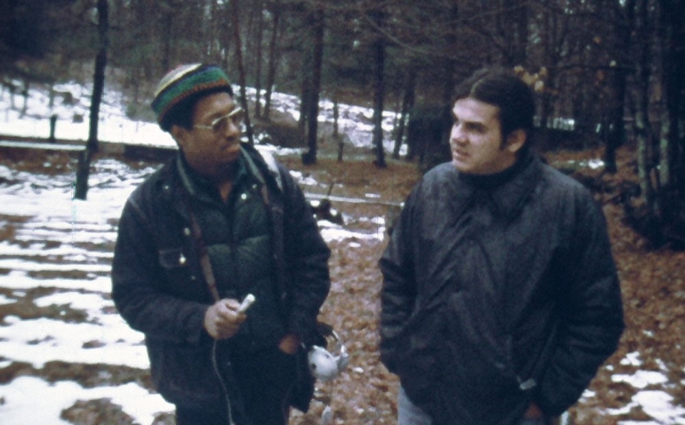 Two men converse in the woods; one holds a microphone to record.