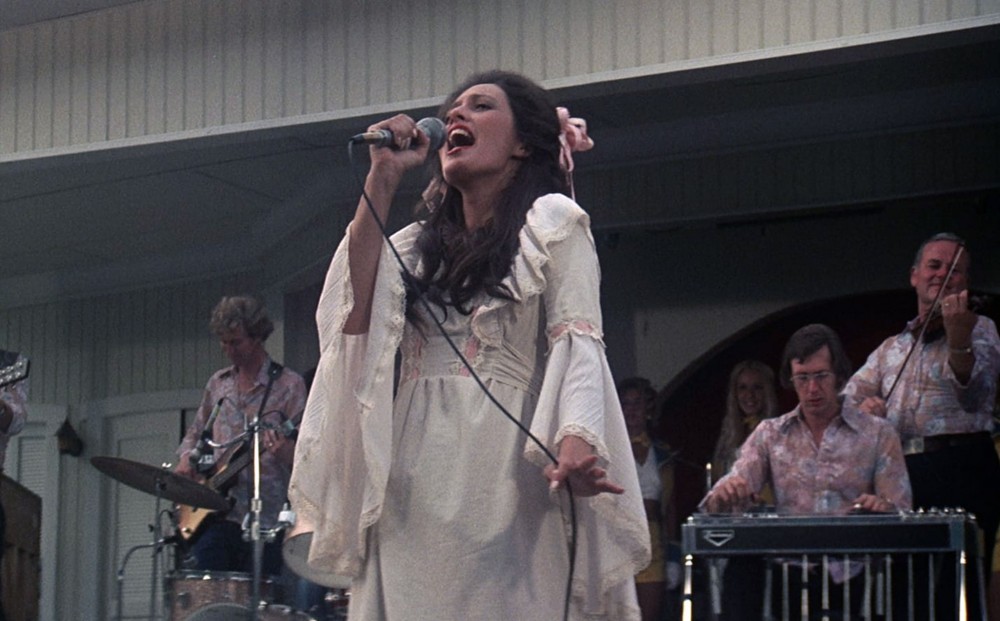 A woman in a long white dress with a pink bow in her long hair sings into a microphone on stage, with a guitarist and a keyboard player behind her.