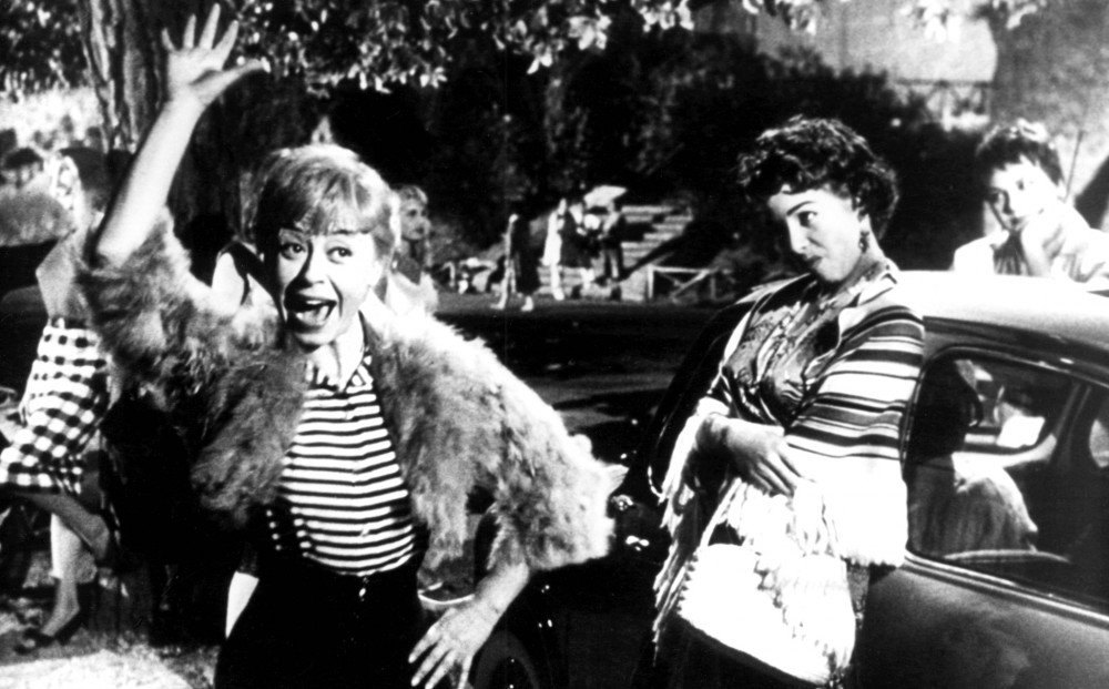 Cabiria poses with her hand on her hip and one arm raised, as though waving hello; two women leaning against a car look at her skeptically.