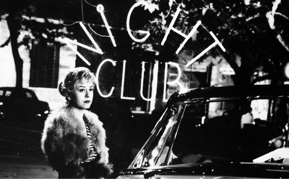 Cabiria stands next to a car, a neon sign reading 