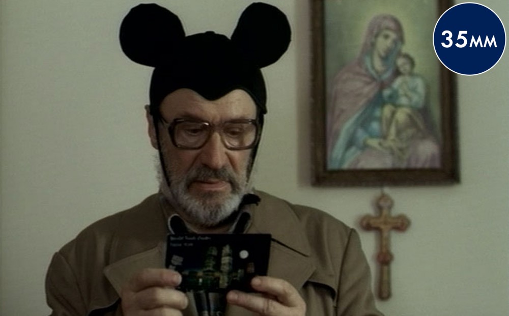 A man wearing a mouse ears hat looks at a postcard; a painting of the Virgin Mary and Jesus is on the wall behind him, with a cross below.