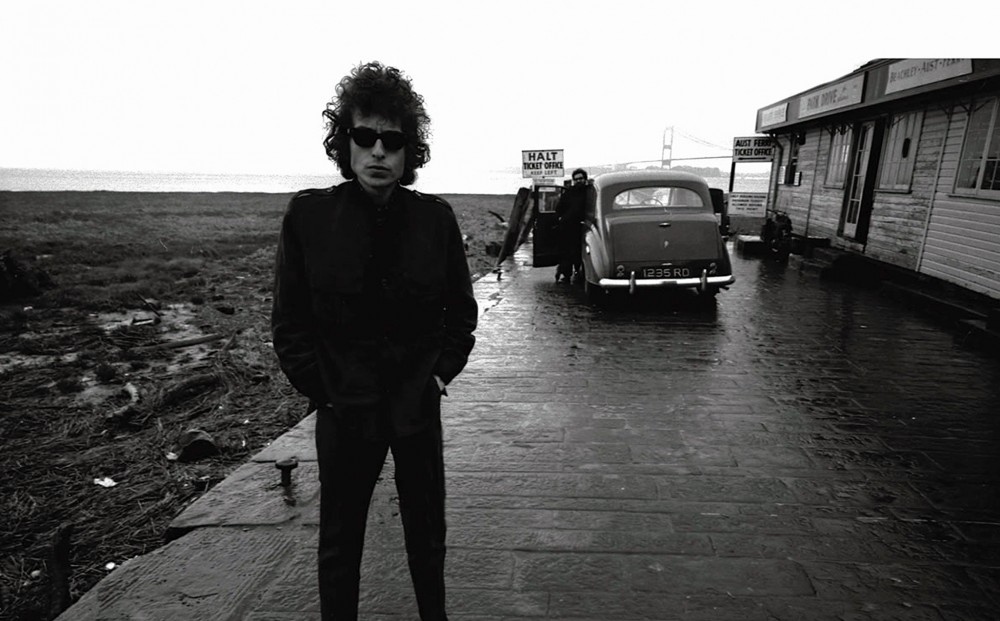 Black and white image of Bob Dylan, facing the camera; a man leans against a car in the background, beyond which you can also see the Golden Gate Bridge.
