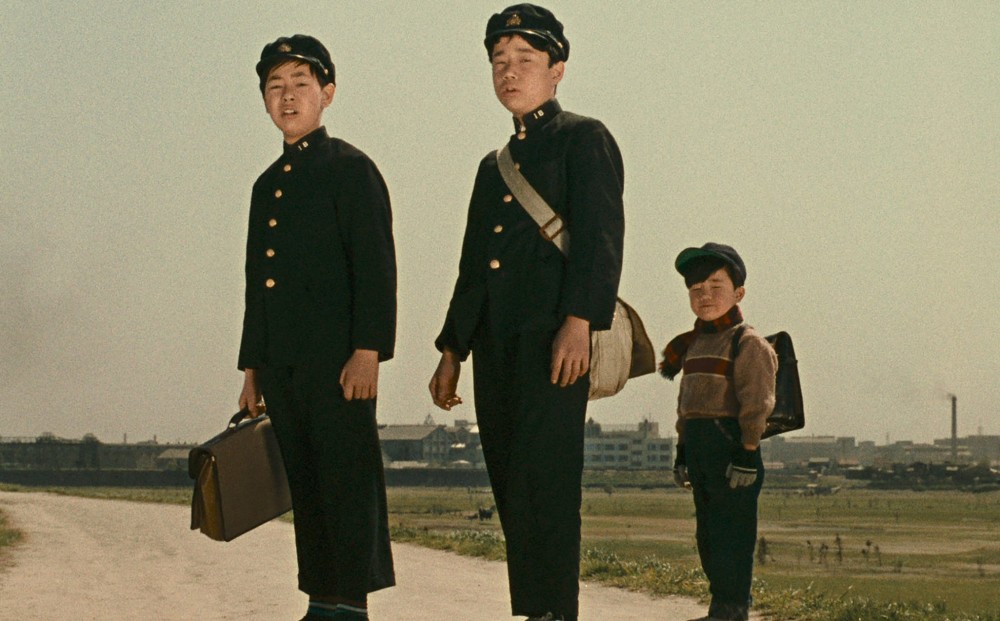 Two young men in uniform and a young boy stand on a gravel path.
