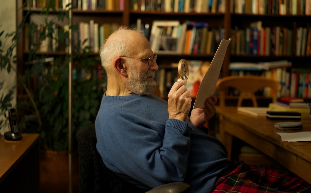 Oliver Sacks sits in a study, reading a paper with the aid of a magnifying glass.