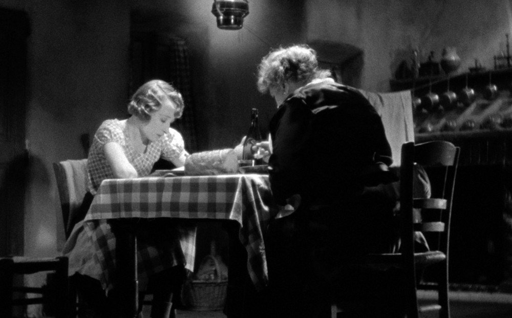Two women sit at a dining room table, both looking down.