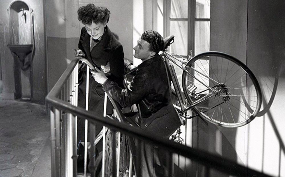 Actor Roger Pigaut carries a bike upstairs on his shoulder while talking to actor Claire Mafféi.