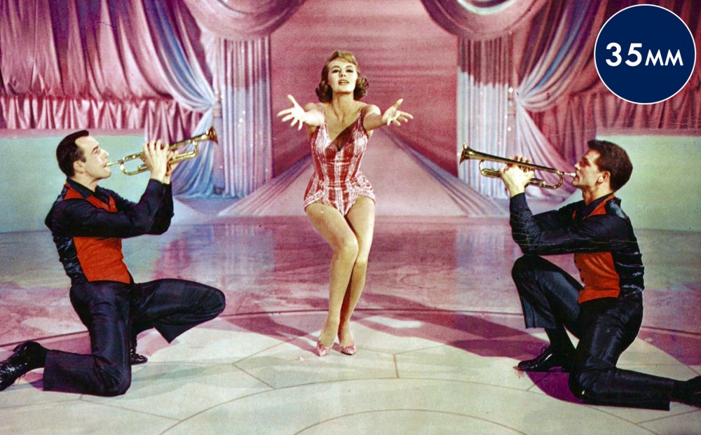 Actor Cyd Charisse stretches her arms out in the middle of a dance number, bordered by two men with trumpets.