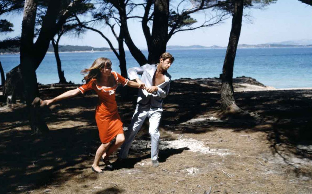 A man holds a woman by the arm, swinging her around in a clearing by the shore.