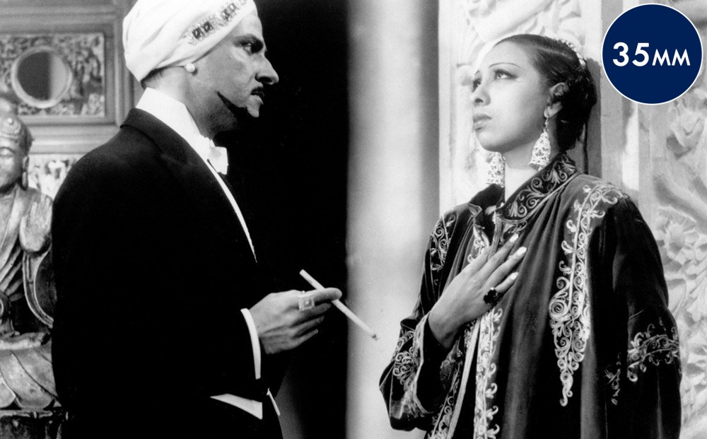 Josephine Baker stands with her hand over her heart, looking melancholy. A man wearing a white turban faces her, holding a long cigarette.