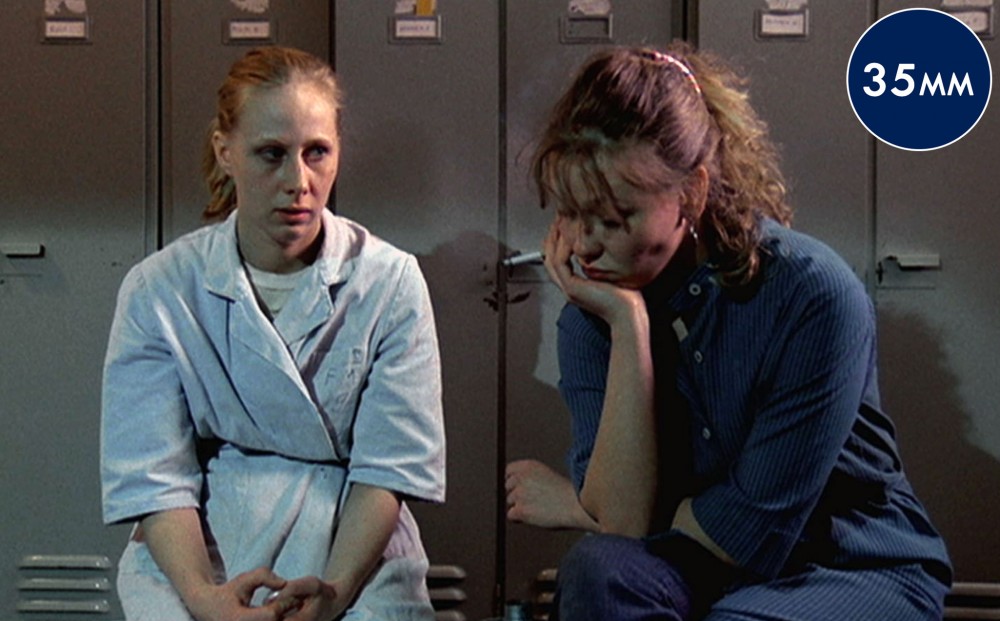 Two women sit in a locker room; one smokes a cigarette.