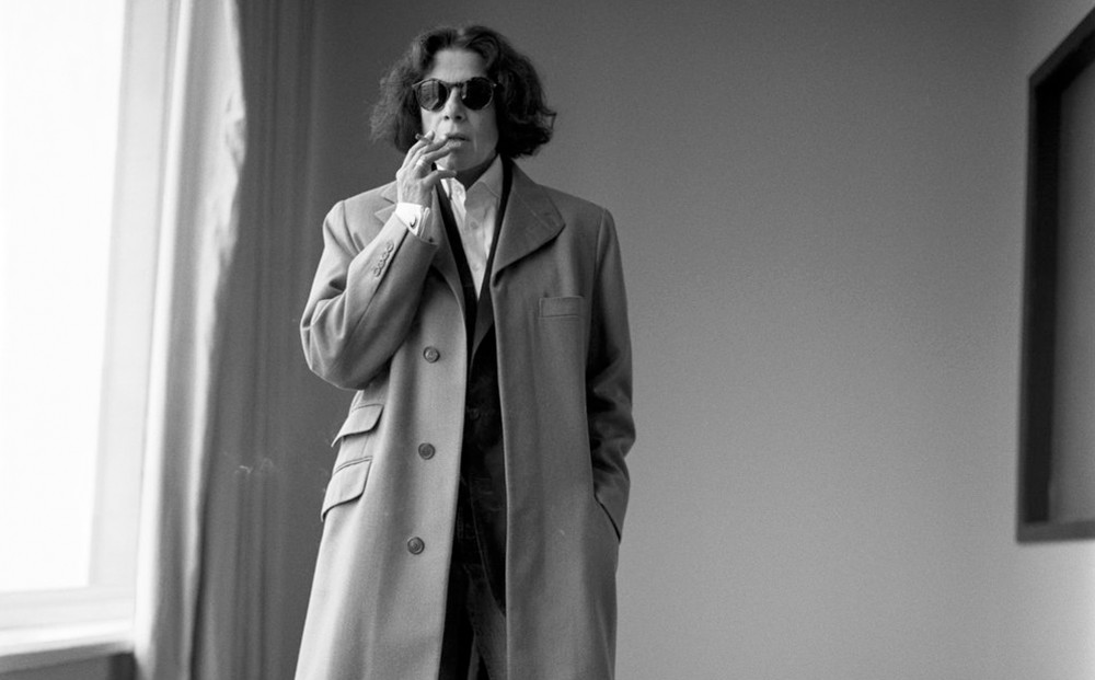 Black and white image of Fran Lebowitz wearing a coat and sunglasses while smoking a cigarette indoors.
