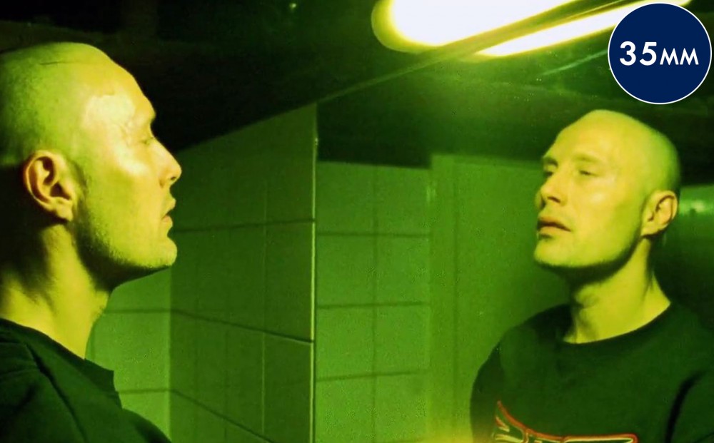 Actor Mads Mikkelsen looks at himself in a bathroom mirror, lit by a green neon light.